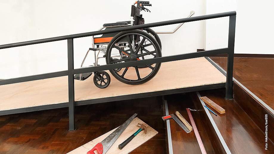Rollstuhl stehend auf einer Rollstuhlrampe. Daneben ist ein Teil der Treppe zu sehen, auf dem Werkzeug liegt.