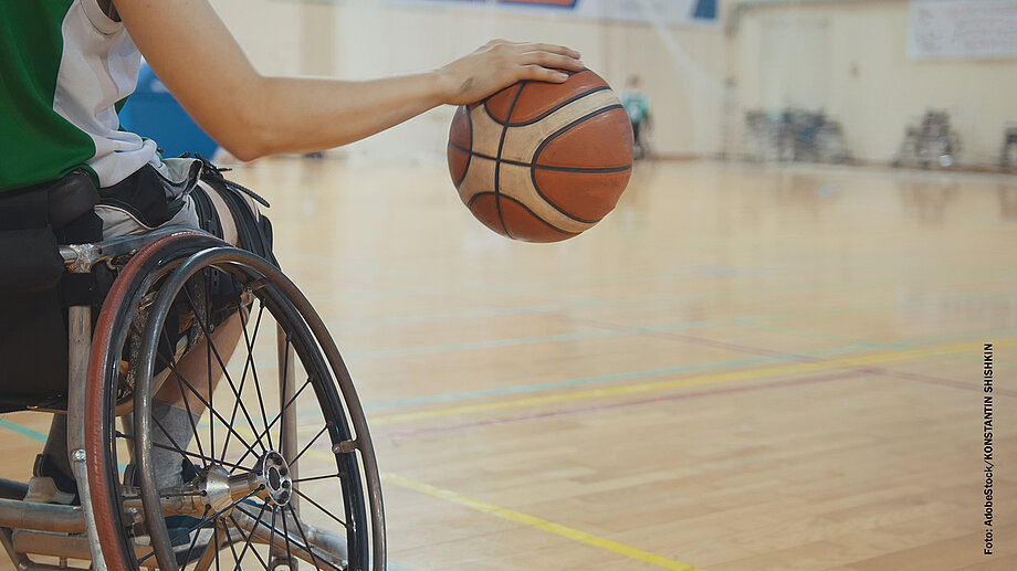 Ansicht von hinten: Person im Rollstuhl dribbelt einen Basketball in einer Sporthalle