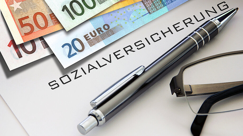 Foto: AdobeStock/Butch Geldscheine, Stift und Brille um das Wort "Sozialversicherung" drapiert